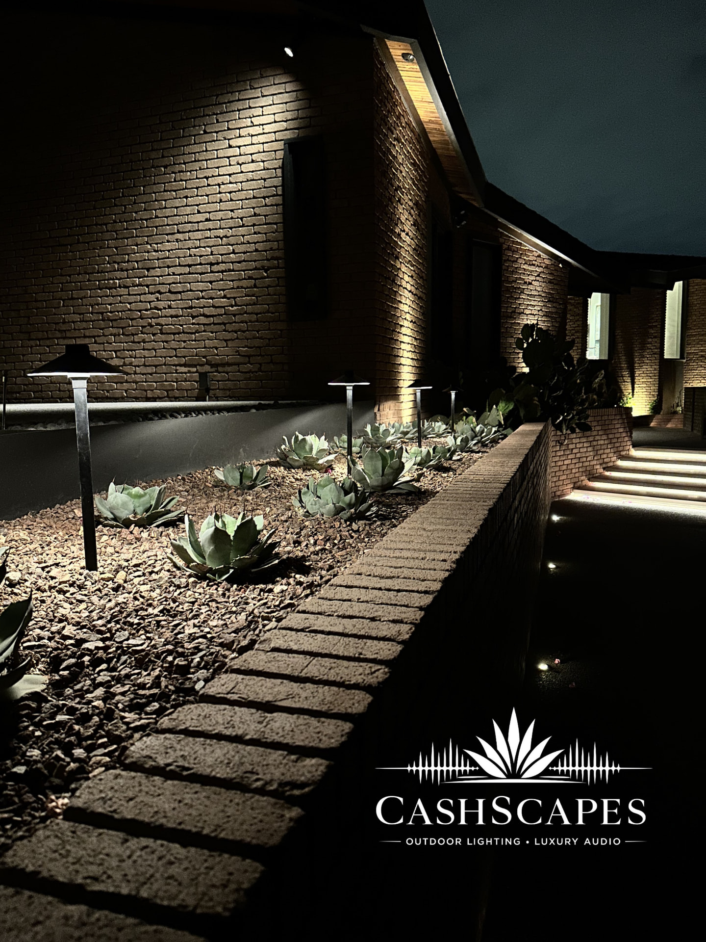 Agave planting bed with path lights and brick wall grazed by warm light